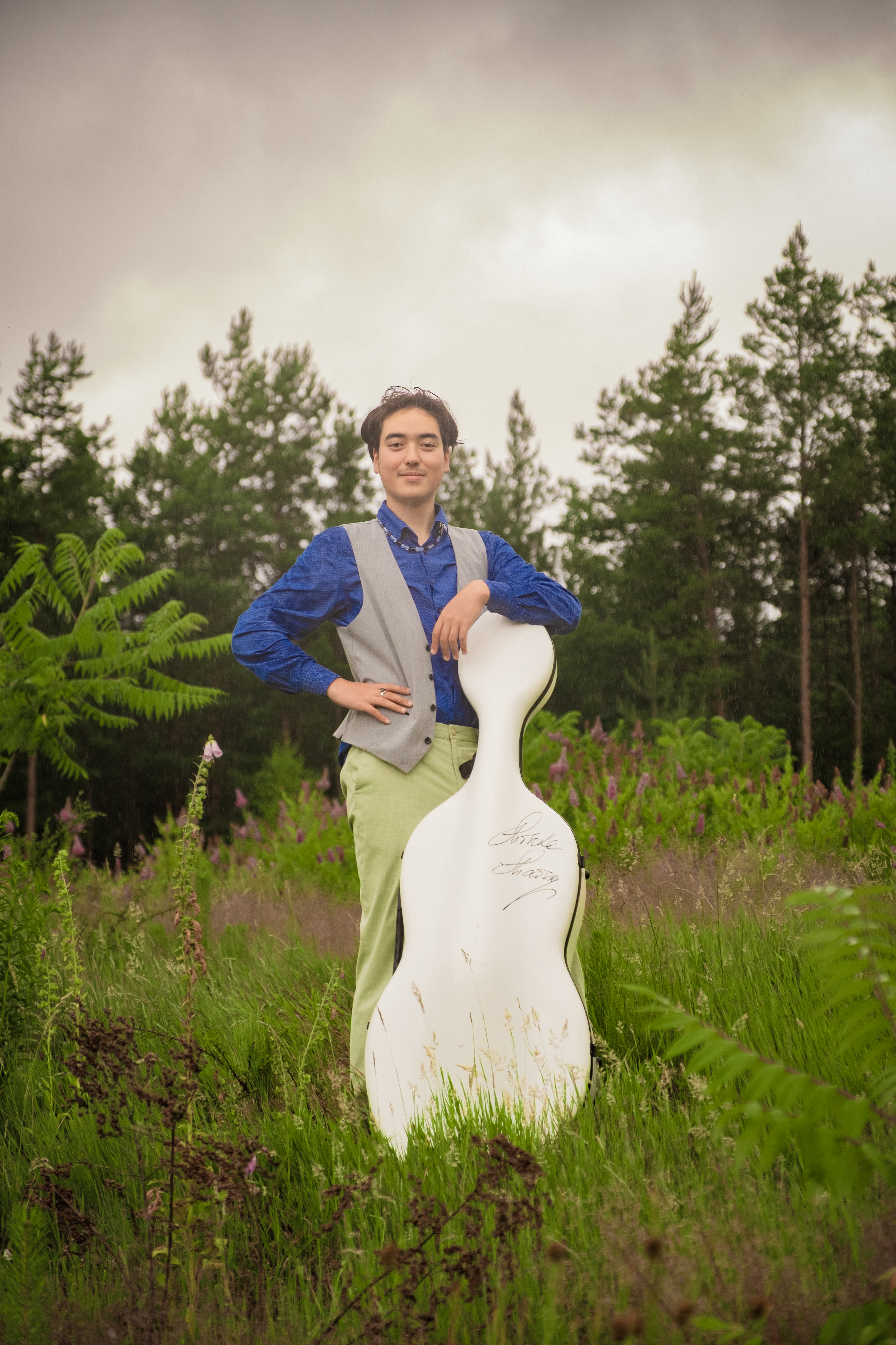 Salomon Held steht neben seinem Cello auf einer Wiese und guckt in die Kamera